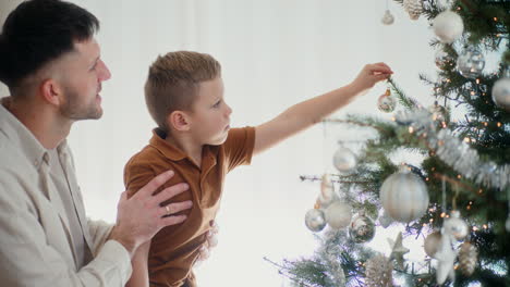 padre con hijo en sus brazos cuelgan bolas de navidad en el árbol