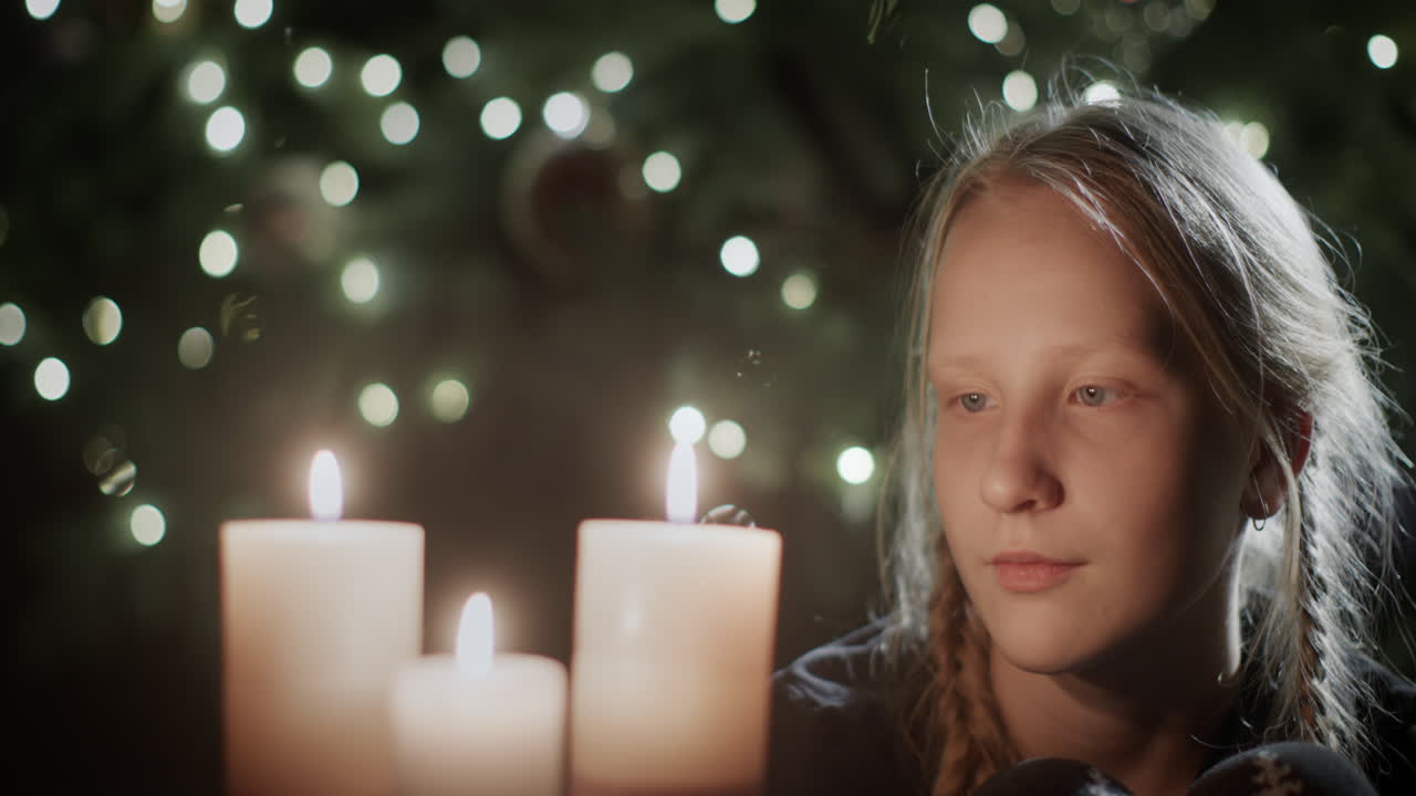 The girl looks at the burning candles. In the background is a beautiful Christmas tree. Children's dreams on Christmas Eve