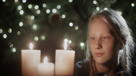 The-girl-looks-at-the-burning-candles.-In-the-background-is-a-beautiful-Christmas-tree.-Children's-dreams-on-Christmas-Eve