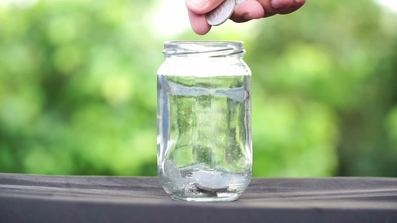 Coins are slowly thrown into a glass jar. Close up shot.