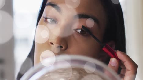 animation of light spots over biracial woman applying mascara