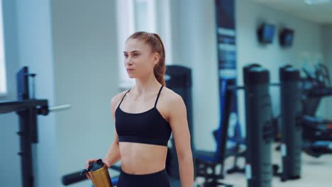 a young woman in black sportswear walks through the gym with a shaker in her hands and drinks water from it. fitness club. blurred background