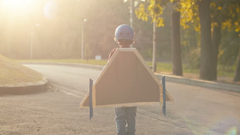 Rückansicht-Eines-Kleinen-Jungen-Mit-Helm-Und-Rotem-Pullover-Mit-Flugzeugflügeln-Aus-Pappe,-Der-An-Einem-Sonnigen-Tag-Im-Freien-Steht-Und-Als-Pilot-Spielt-1