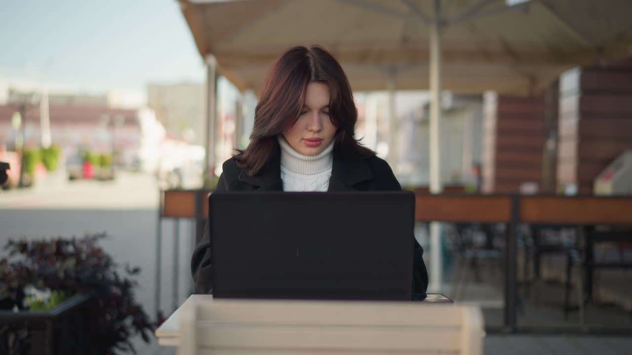 Lady working on laptop in urban setting, wearing stylish black coat, with decorative black pillar nearby, blurred red brick building in background, and blurred figure with greenery in the distance
