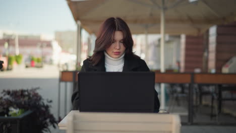 lady working on laptop in urban setting, wearing stylish black coat, with decorative black pillar nearby, blurred red brick building in background, and blurred figure with greenery in the distance