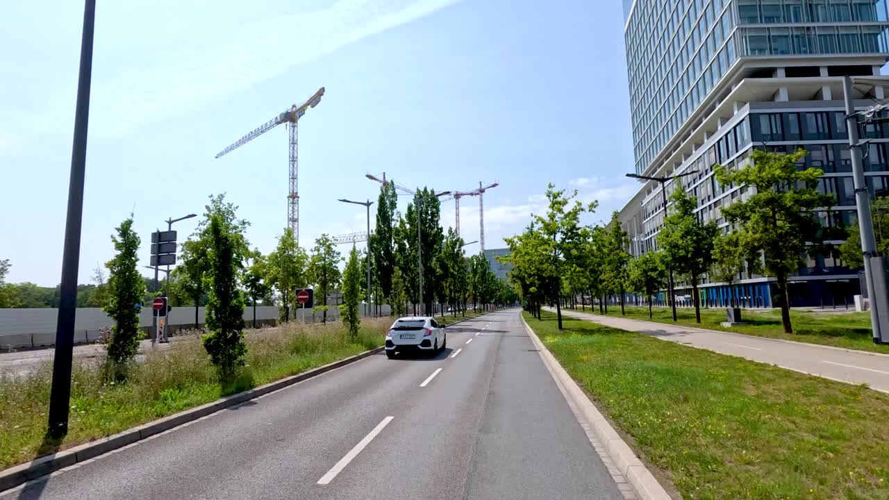 White car drives down tree-lined city street with modern glass buildings in bright daylight