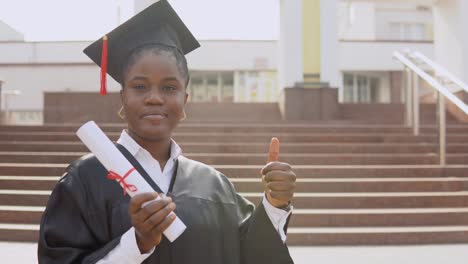 joven graduada afroamericana de pie frente a la cámara con un diploma y libros en las manos. la estudiante está vestida con una túnica negra y un sombrero de maestro cuadrado y está de pie afuera.