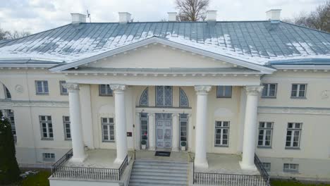 aerial drone footage slowly rising higher and ascending in front of saku manor or saku mõis during a winter day with green grass and leafless bare trees around. large white building with pillars.
