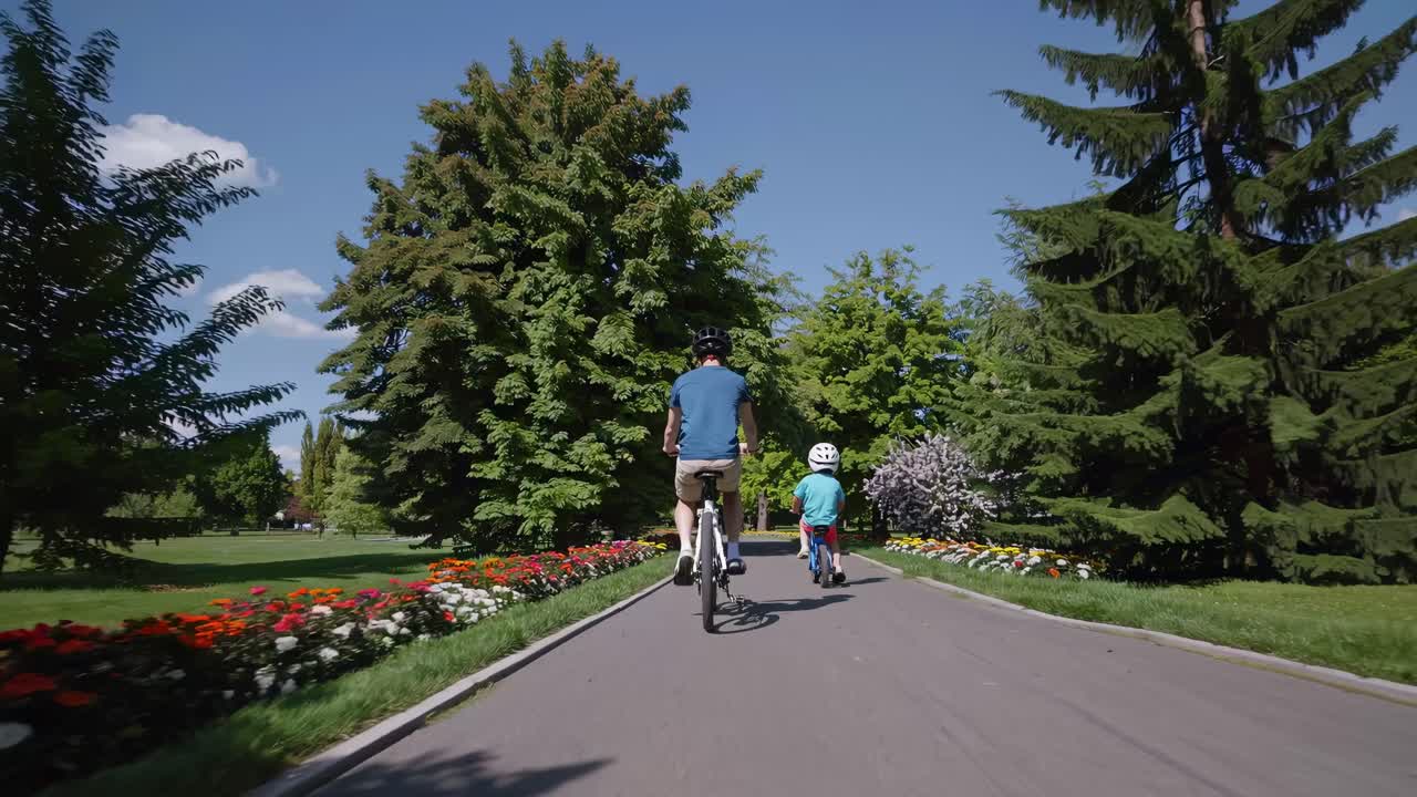 A video captures a rear-view angle of a father and child cycling on a park path, surrounded by lush