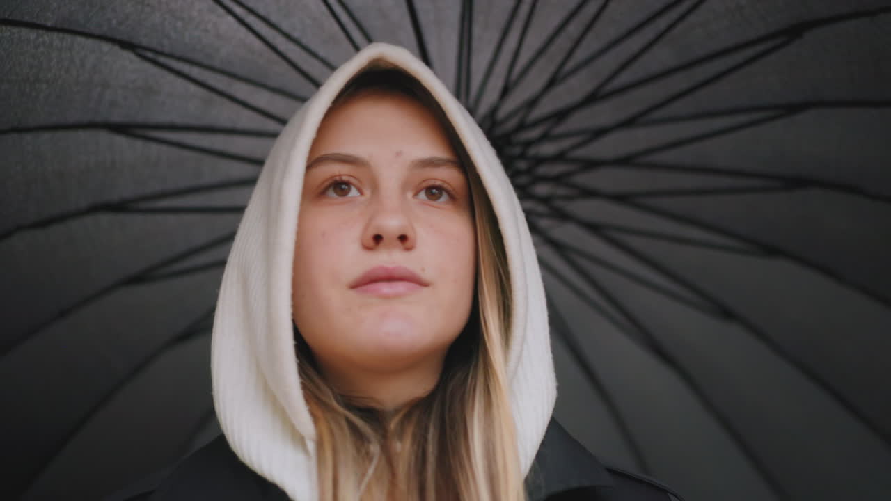 Portrait of young woman in white hood standing under large black umbrella, looking ahead with calm thoughtful expression, outdoor rainy day scene with moody atmosphere and introspective urban vibe