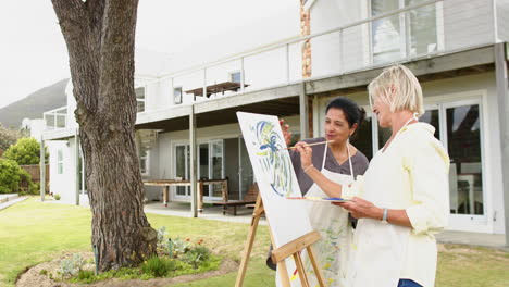 Two-happy-diverse-senior-women-painting-picture-and-laughing-in-sunny-garden,-slow-motion