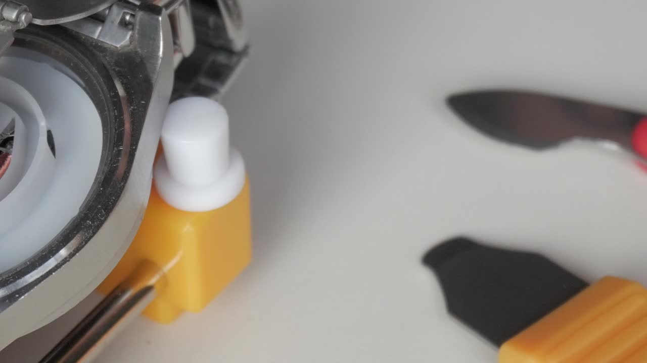 Macro view of a watch repair tool in use, showing metal components and yellow plastic parts, panning right