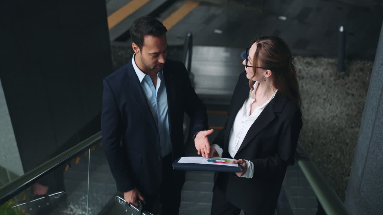 Une femme cadre dirigeante souriante montre un diagramme circulaire financier coloré à un collègue masculin tout en montant un escalator dans un espace d’entreprise moderne