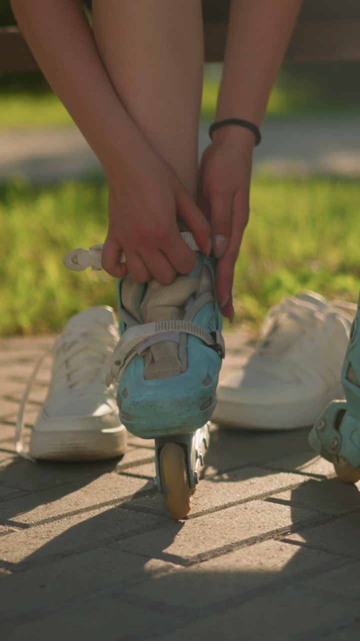 벤치에 앉아 있는 개인의 다리 시각. 오른손에 검은색 팔를 가진 시안 롤러 스케이트의 줄을 조정하는 동안  스니커가 보행길에 뒤에 고 있습니다.