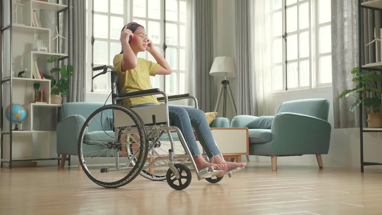 Asian Kid Girl Sitting In A Wheelchair Listening To Music With Headphones And Dancing In Living Room