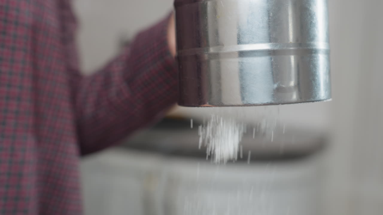 primer plano de una persona tamizando harina de maíz a través de un filtro de copa de acero inoxidable, las partículas finas de harina caen suavemente, creando una textura suave. el fondo presenta un entorno de cocina con decoración moderna