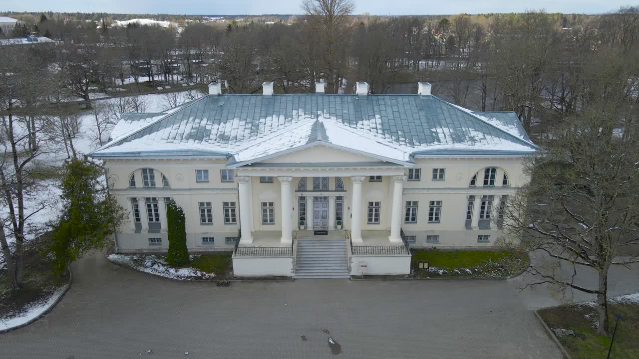 Gorgeous aerial drone footage view descending and flying lower in front of a large white and historic Saku manor mansion or Saku Mõis during a winter cloudy day, with leafless trees and snow around it