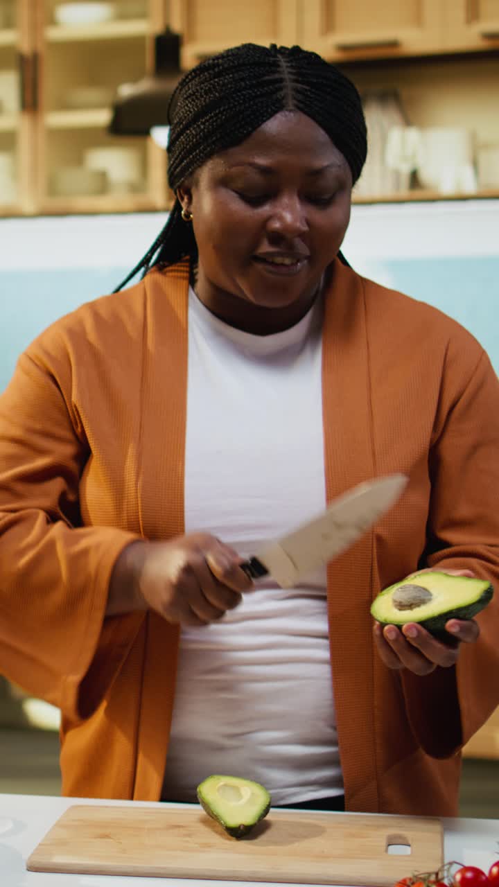 Video verticale di una donna afroamericana che registra il taglio di un avocado in un vlog di cucina sul bancone della cucina