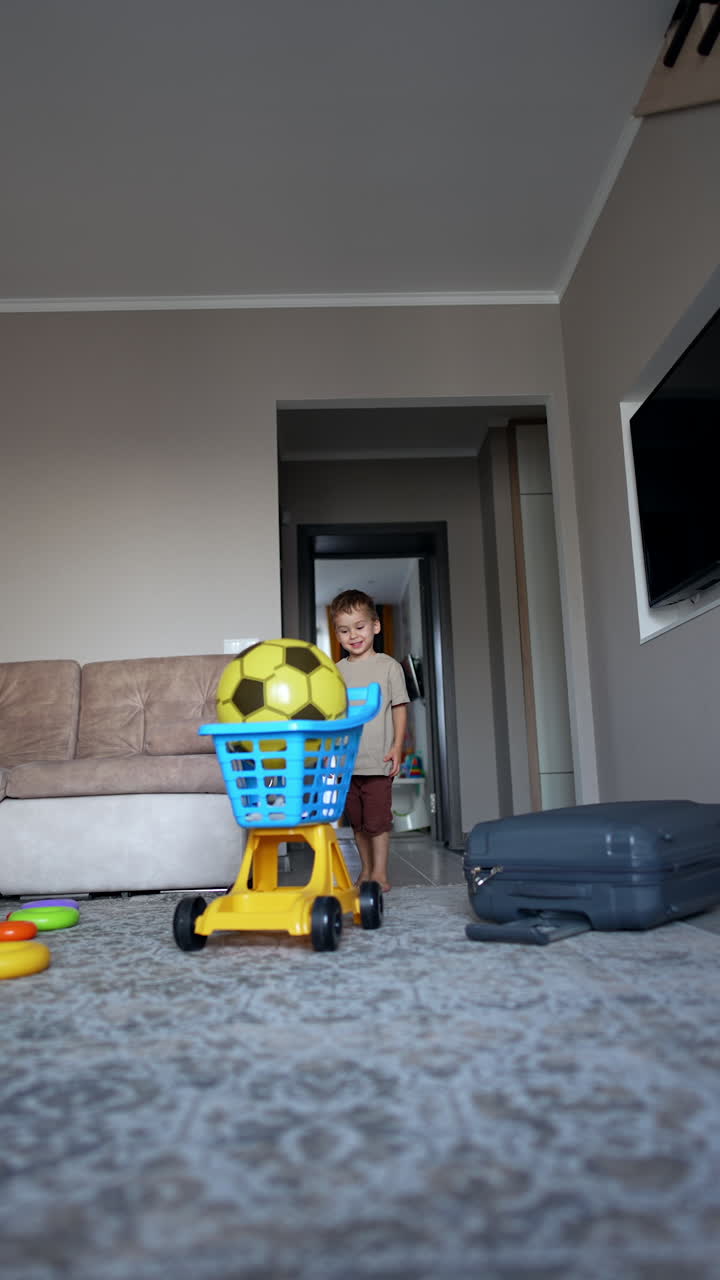 Adorable toddler boy pushes a cart with a big yellow ball. A big grey suitcase lies on the floor. Vertical video.
