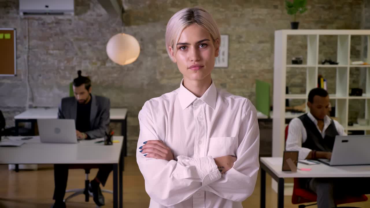 Young pretty businesswoman is standing and crossing her arms in office, colleagues are working with laptops, work concept