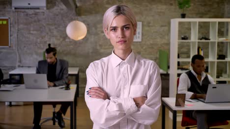 young pretty businesswoman is standing and crossing her arms in office, colleagues are working with laptops, work concept