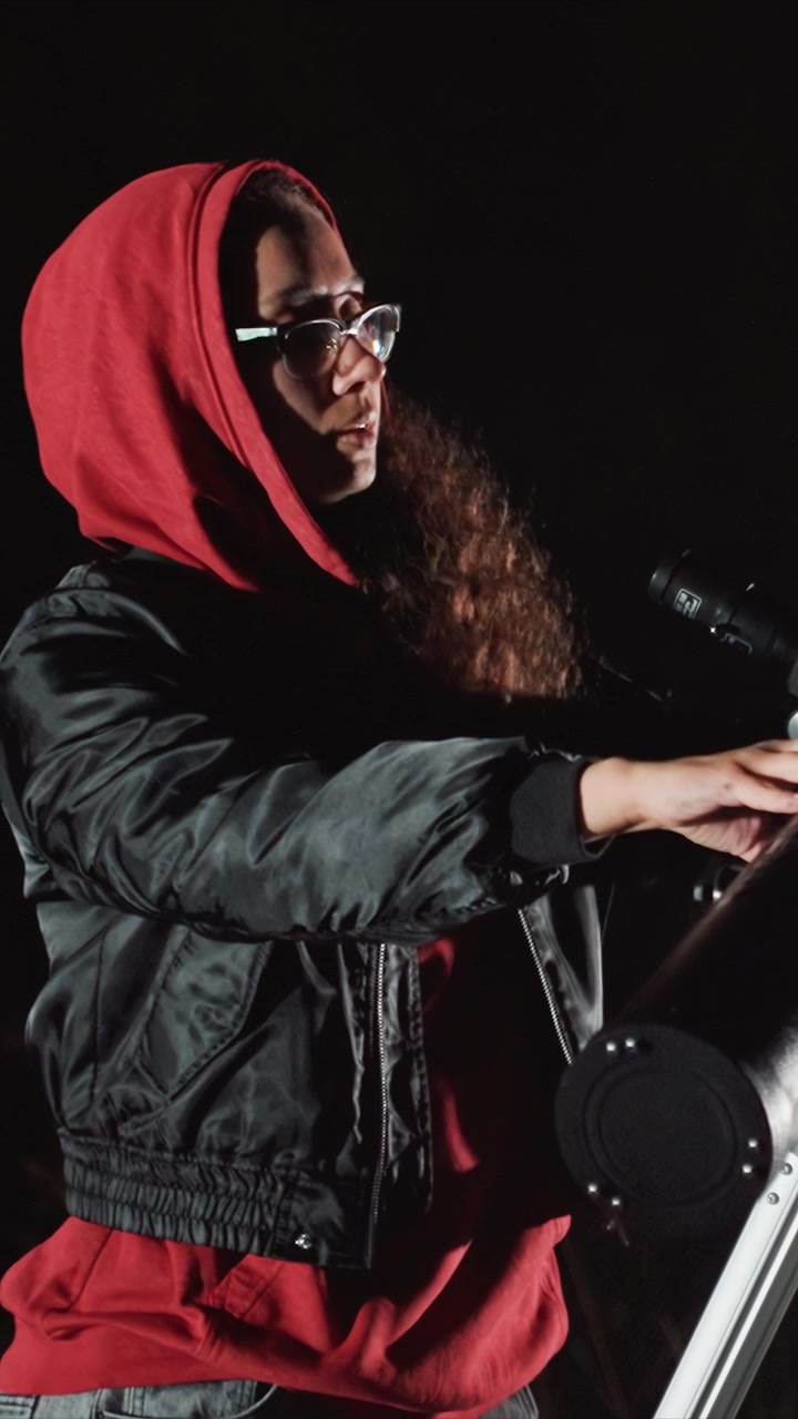 Woman in red hoodie and black jacket stands before microphone. Female adjusts sizable viewing apparatus employing. Shadowy scene includes visible parts of recording tools