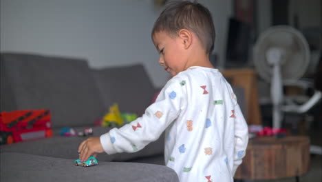 joven hispano en pijama blanco jugando con un pequeño juguete de coche azul en un sofá gris