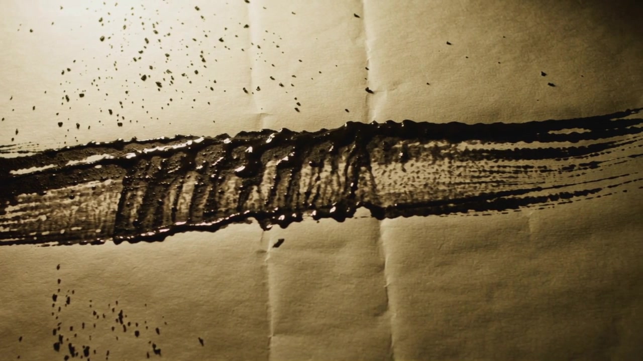 Dragging brush starting left edge, pulling ink across brown paper on table to create stroke