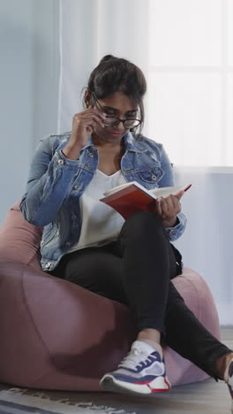 relaxed indian woman reads book sitting on bean bag chair in living room against bright window. young lady reader enjoys captivating tale at home