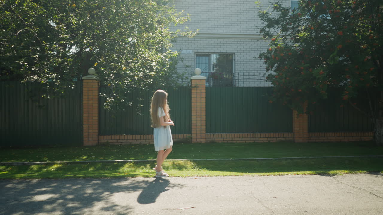 Side view of young girl walking calmly by road edge as sunlight creates glowing effect through trees, with green fence, brick pillars, and soft shadows cast on pavement in peaceful suburban setting