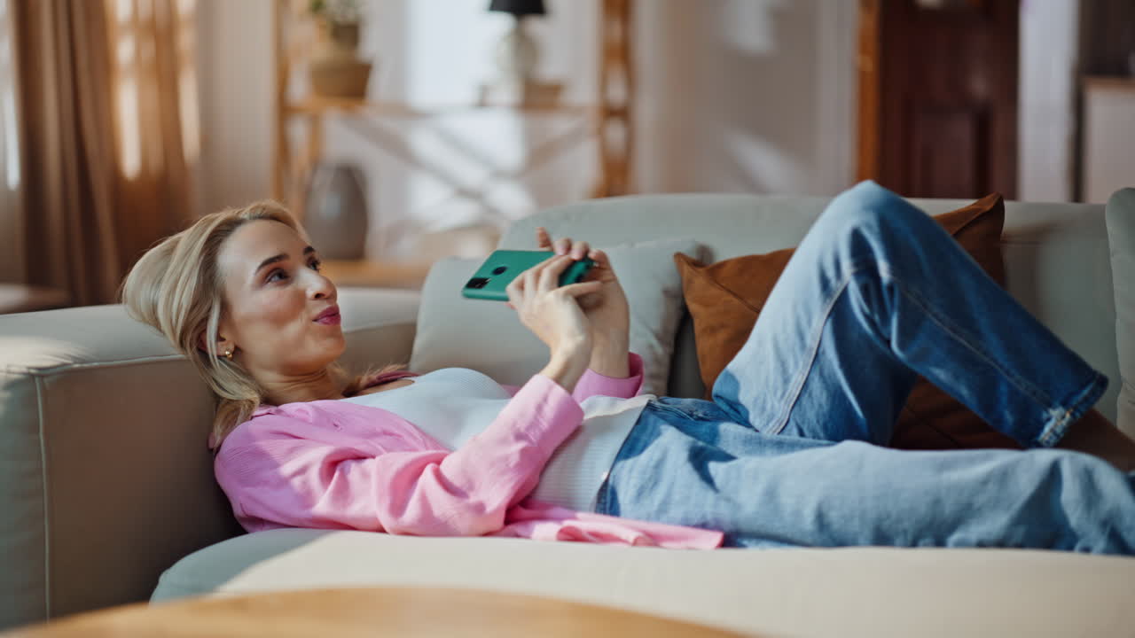 Chilling blonde watching cellphone lying sofa. Smiling woman looking smartphone