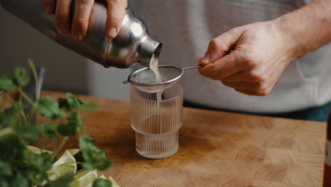 Young-male-pouring-a-cocktail-from-mixer-through-sieve-into-modern-drinking-glass-in-the-kitchen-at-home