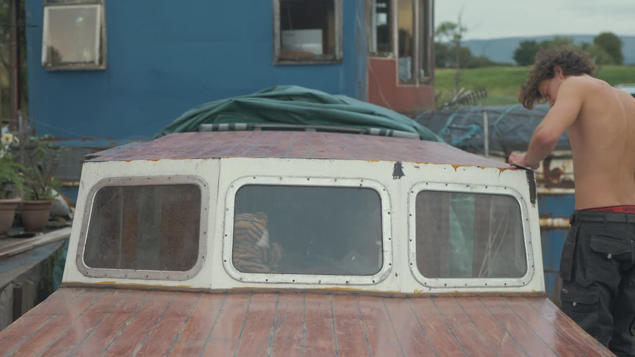 joven carpintero en topless entra al marco y comienza a lijar la cabina del techo del barco usando papel de lija