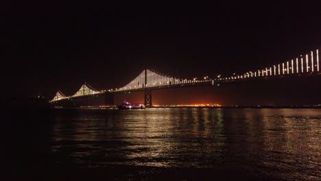 Gimbal-static-shot-of-a-ferry-passing-under-the-Bay-Bridge-at-night-in-San-Francisco,-California