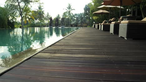 Zeitlupen-Dollyaufnahme-Eines-Pools-Mit-Blick-Auf-Den-Holzweg-Und-Einer-Liegewiese-Mit-Bäumen-Und-Palmen-Im-Hintergrund-Auf-Bali,-Indonesien