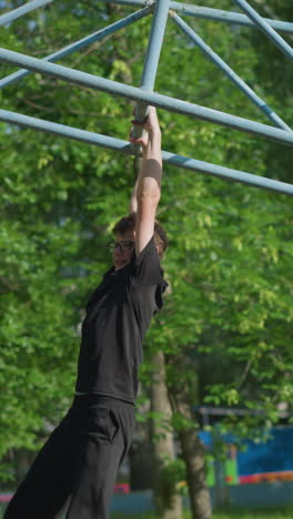 A young boy wearing a black outfit and glasses swings slowly on outdoor equipment, with lush green trees and a fence in the background