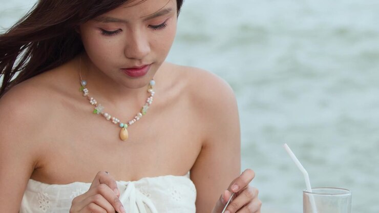 Ripresa a figura intera di una donna asiatica con un abito senza spalline che mangia all'aperto vicino al mare