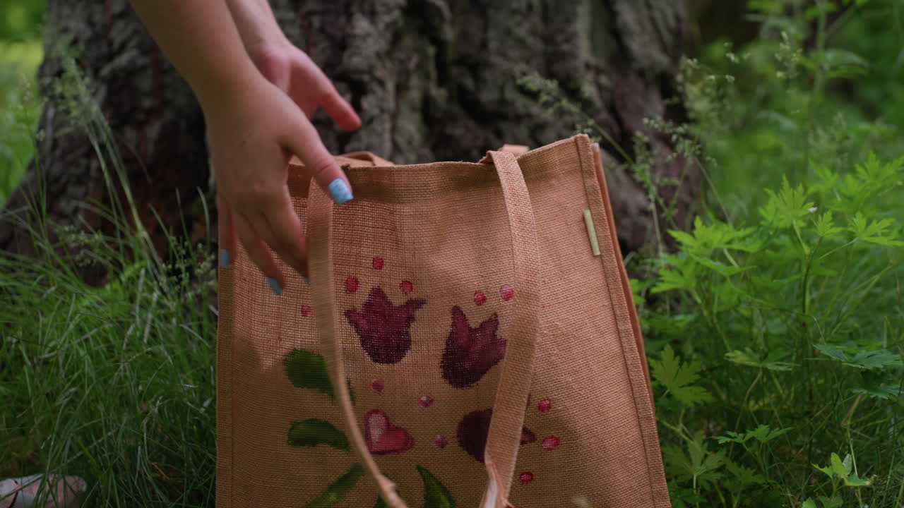 Lower angle view of person returning blanket into eco bag beside tree trunk on green grass, then carrying bag and walking away under soft daylight