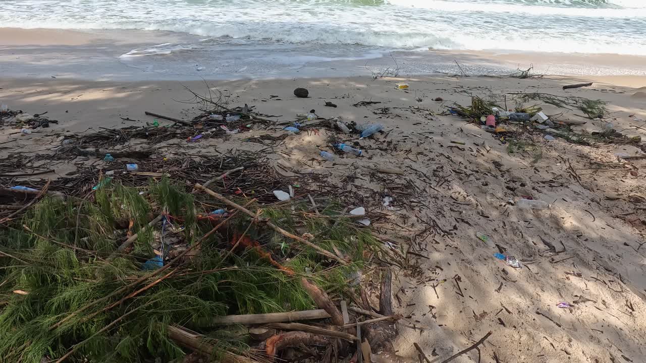 A sandy beach in Ko Phayam, Thailand, shows exposed tree roots, scattered plastic debris, and driftwood under bright daylight with steady camera movement
