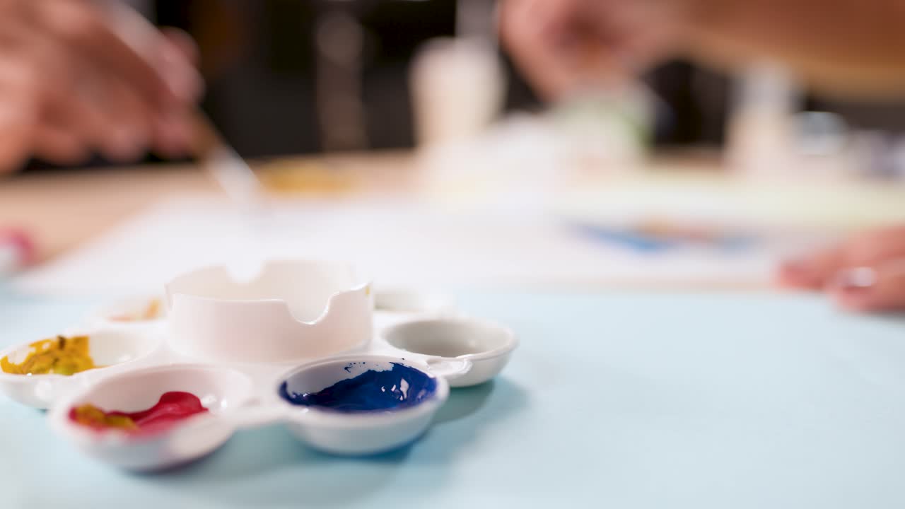 Kids' hands mix paint with brushes on palette in bright, indoor craft workspace, shallow focus