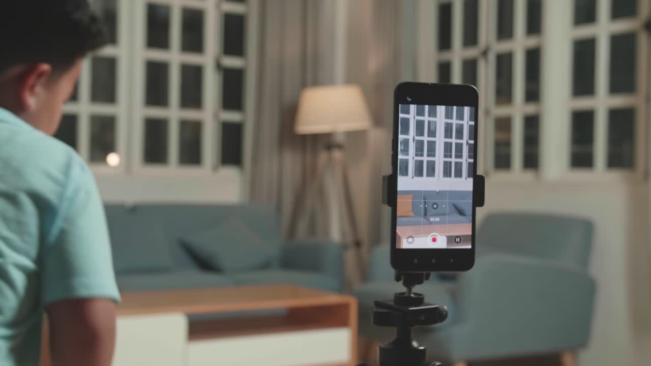 Display Smartphone Of Asian Little Boy Shows Trick With Disappearing On Mobile Phone Camera While Recording Video Content For Social Networks In Cozy Room