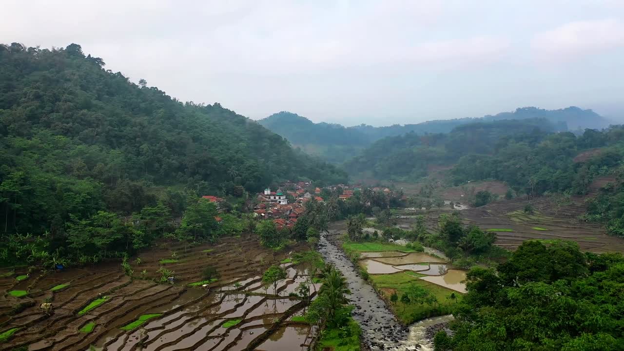 Drone captures terraced rice fields river and dense green hills with small village nestled in valley in Nanggung, Indonesia under cloudy sky showing peaceful rural lifestyle and natural beauty