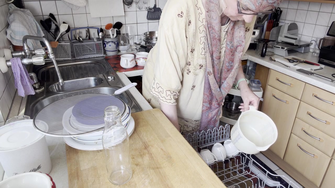 A static kitchen shot showing a woman attempting to place a large mixing bowl into the dishwasher, returning it after checking the fit, then loading a smaller plastic container into the upper rack.