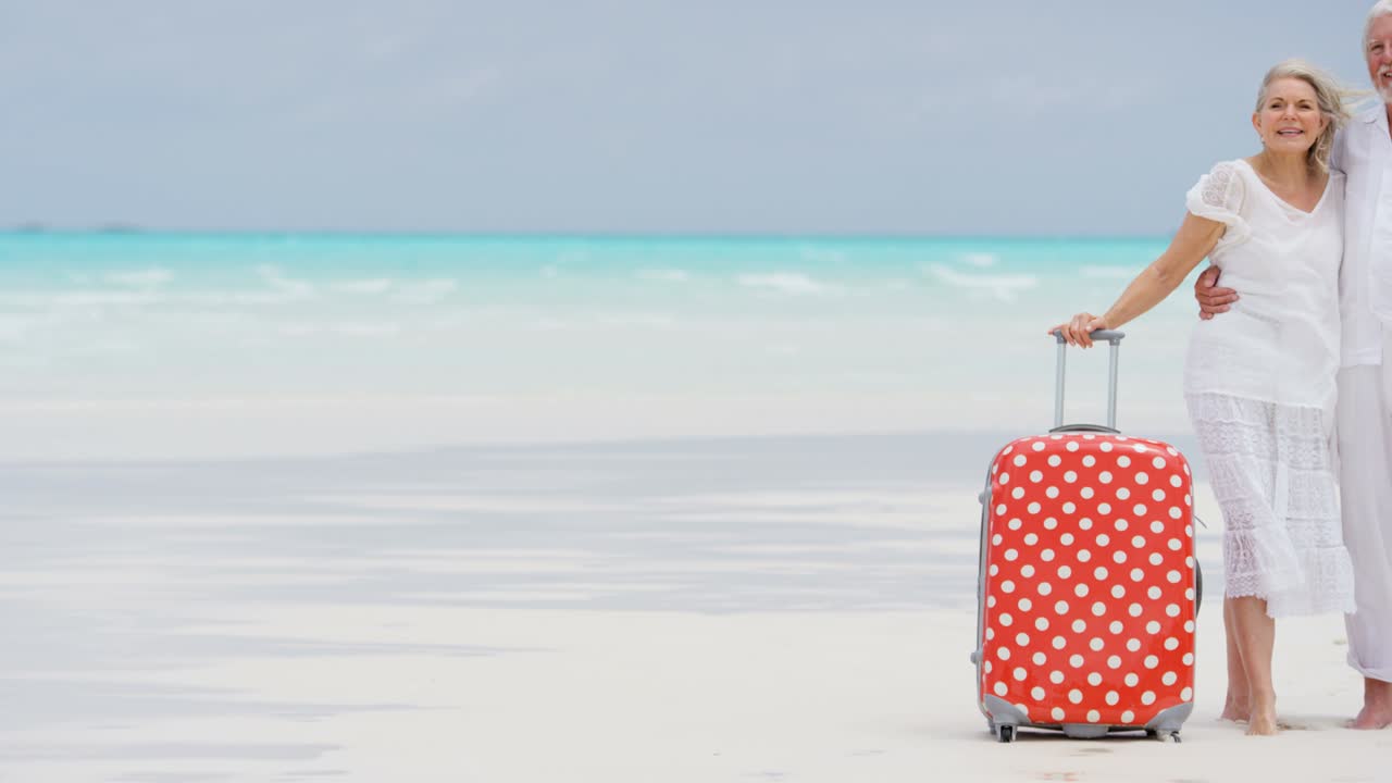 старшая кавказская пара стоит на пляже с чемоданом