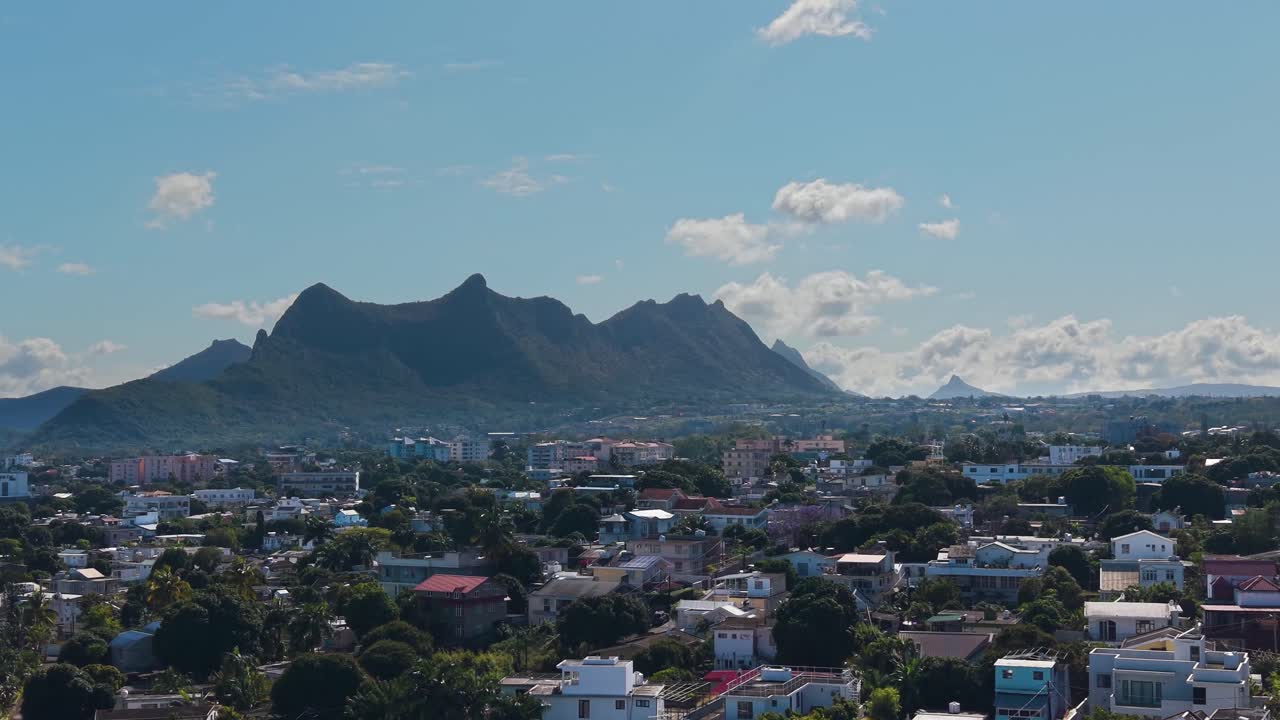 Aerial view of Beau Bassin–Rose Hill, Mauritius, showcasing a tropical suburban landscape with colorful houses, lush greenery, and the majestic mountain range rising in the background under a blue sky