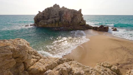 beautiful-mediterranean-beach-in-spain,-overlooking-turquoise-and-blue-waters,-with-a-rock-in-the-middle-of-the-sea