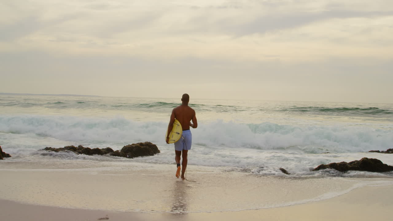 vista trasera de un surfista afroamericano corriendo con una tabla de surf en la playa 4k