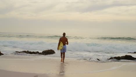 Rückansicht-Eines-Afroamerikanischen-Männlichen-Surfers,-Der-Mit-Surfbrett-Am-Strand-Läuft-4k