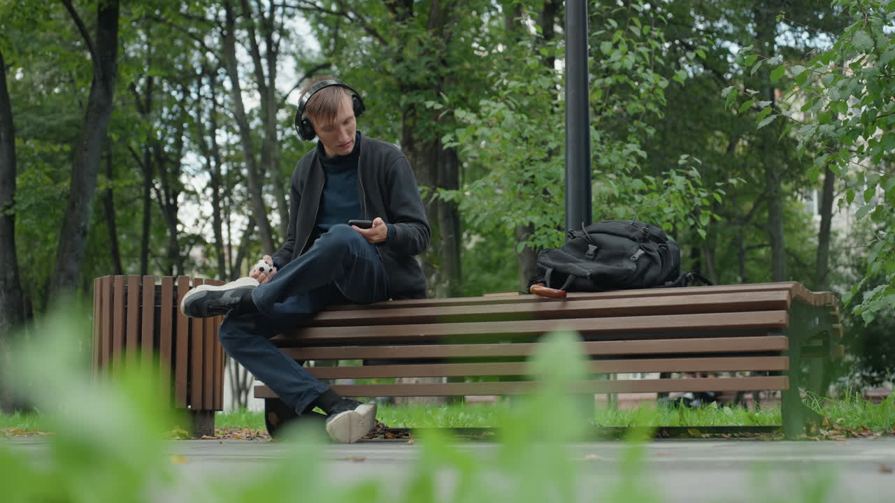 Osamělý muž sedí na lavičce v parku, má na sobě sluchátka a prohlíží si smartphone, batoh vedle něj, zelené stromy a zamračené světlo vytvářejí reflexivní klid, ležérní bunda, jemný vánek, pomalé kývání hlavou