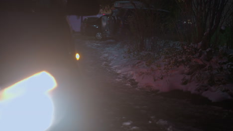 illuminated car headlamp casting a bright beam on icy road surrounded by snow and scattered vegetation, with blurred parked cars and faint buildings in the background